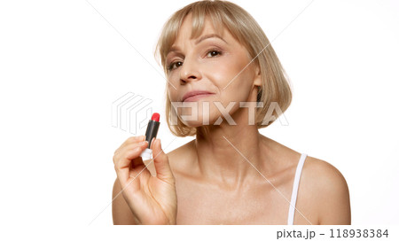 Mature woman holds lipstick confidently, ready to apply it, showing self-expression and beauty confidence against white background. Mature woman holds lipstick confidently, ready to apply it, showing self-expression and beauty confidence against white background. 118938384
