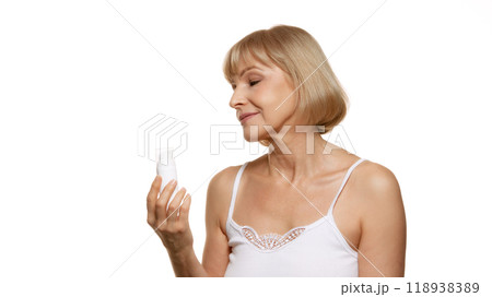 Inspecting her skincare product with calm gaze, beautiful woman chooses her self-care rituals with purpose against white studio background. 118938389