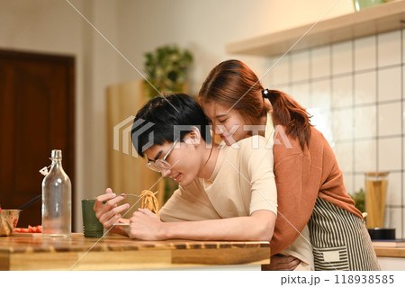 Pretty young woman embracing her boyfriend lovingly from behind while he eating pasta in the kitchen Pretty young woman embracing her boyfriend lovingly from behind while he eating pasta in the kitchen 118938585