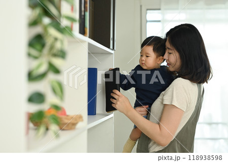 Beautiful mother with a baby boy standing next to bookshelf at home. Childhood and nurturing care concept 118938598