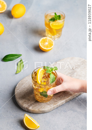 A hand takes a glass of iced tea with lemon and mint against a gray background with citrus fruits. 118939027