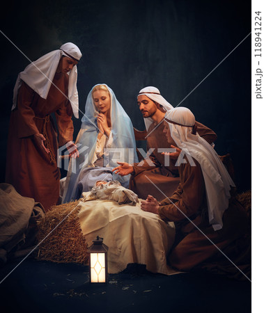 Tranquil nativity scene, with Mary and Joseph in prayer over baby Jesus. Shepherds showing reverence, reflecting holiness of sacred night. 118941224