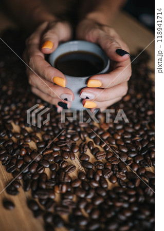 A warm cup of coffee held in woman hand with colorful nails surrounded by fresh coffee beans on a A warm cup of coffee held in woman hand with colorful nails surrounded by fresh coffee beans on a 118941714