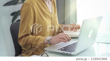 Unrecognizable professional woman is making notes and working with laptop in modern office. Yellow blouse is passing to adult businesswoman. Business concept Unrecognizable professional woman is making notes and working with laptop in modern office. Yellow blouse is passing to adult businesswoman. Business concept 118941900