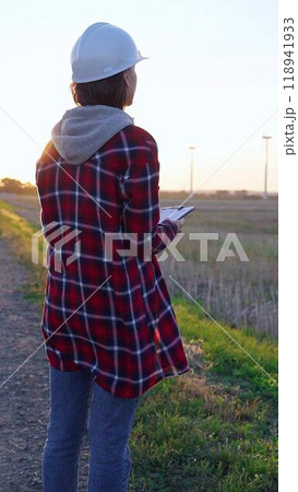 Woman engineer wearing a white protective helmet is taking notes with a clipboard in a field with wind turbines. Clean energy and engineering concept Woman engineer wearing a white protective helmet is taking notes with a clipboard in a field with wind turbines. Clean energy and engineering concept 118941933