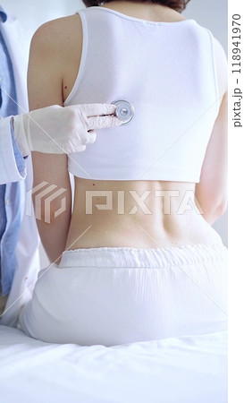 Doctor and patient. Physician wearing white medical gloves is using stethoscope examining woman lungs from the back side in clinic. Medicine and health care 118941970