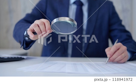 Businessman is carefully examining financial documents with a magnifying glass, highlighting the importance of thorough analysis in business. Audit and taxes 118941972