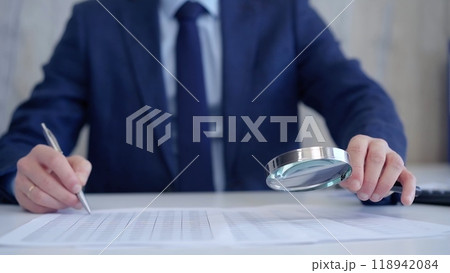 Businessman is carefully examining financial documents with a magnifying glass, highlighting the importance of thorough analysis in business. Audit and taxes 118942084