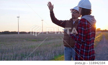Man and woman, both engineers wearing white protective helmets, are checking and discussing something in a field with wind turbines as the sun sets. Clean energy and engineering concept 118942104