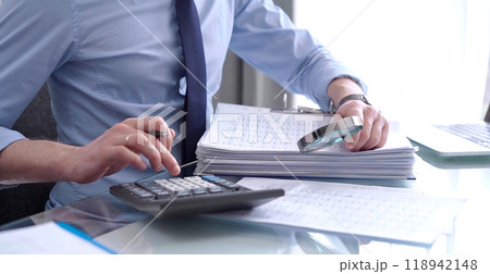 Businessman in blue short is using calculator and reviewing ring folder of financial documents with magnifying glass at desk in modern office. Audit and taxes in business concept 118942148