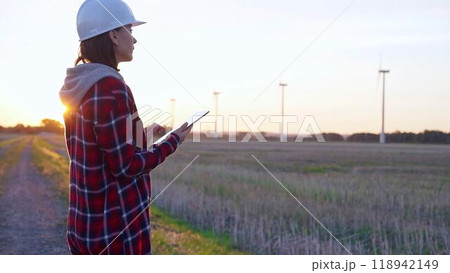 Woman engineer wearing a white protective helmet is taking notes with a tablet computer on a field with wind turbines, as the sun sets. Clean energy and engineering concept 118942149