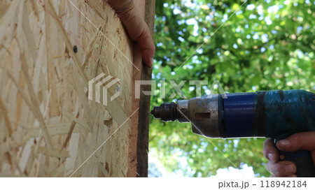 old drill makes holes in the surface of an outbuilding made of wood and chipboard sheets, close-up, working with a drill in domestic conditions old drill makes holes in the surface of an outbuilding made of wood and chipboard sheets, close-up, working with a drill in domestic conditions 118942184