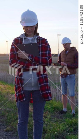 Woman engineer is standing straight and taking notes with a clipboard in a field with wind turbines, as the sun sets. Clean energy and engineering concept Woman engineer is standing straight and taking notes with a clipboard in a field with wind turbines, as the sun sets. Clean energy and engineering concept 118942189