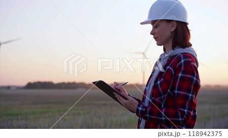 Woman engineer taking notes on a clipboard on a field with wind turbines, as the sun sets. Clean energy and engineering concept 118942375