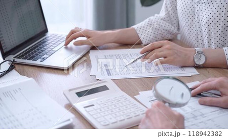 Female bookkeeper or auditor is working on finances papers with a colleague, using magnificent glass, calculator and laptop. Audit and taxes 118942382