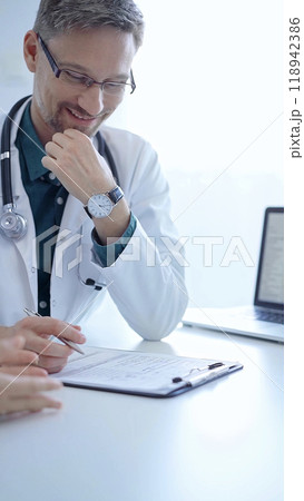 Doctor and a patient. The physician, wearing a white medical coat over a green shirt, gesturing with his hands during a consultation in the clinic. Medicine concept 118942386