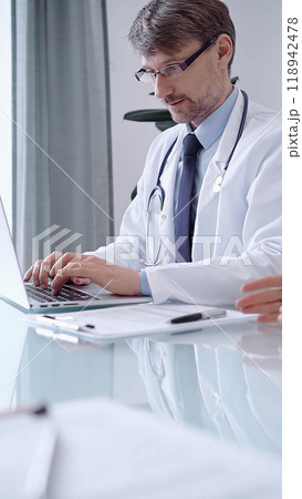 Male physician in white coat focused on laptop data in a modern clinic office while analyzing patient information on laptop. Medicine concept Male physician in white coat focused on laptop data in a modern clinic office while analyzing patient information on laptop. Medicine concept 118942478