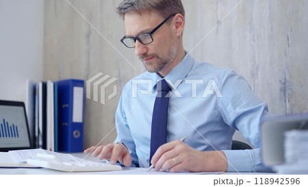 Male accountant with blue t-shirt concentrating while calculating costs and taking notes at his desk. Taxes, audit in business 118942596