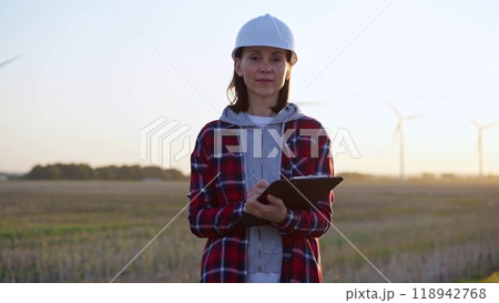 Female engineer is standing straight and keeping clipboard on a field with wind turbines, as the sun sets. Concept of clean energy and engineering audit Female engineer is standing straight and keeping clipboard on a field with wind turbines, as the sun sets. Concept of clean energy and engineering audit 118942768