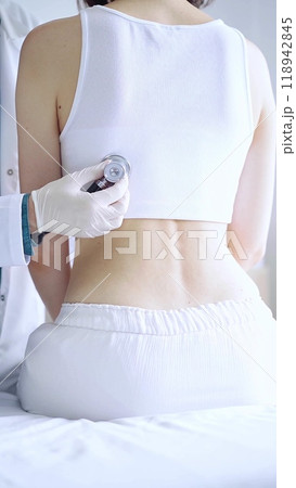 Doctor and patient. Physician wearing white medical gloves is using stethoscope examining woman lungs from the back side in clinic. Medicine and health care Doctor and patient. Physician wearing white medical gloves is using stethoscope examining woman lungs from the back side in clinic. Medicine and health care 118942845