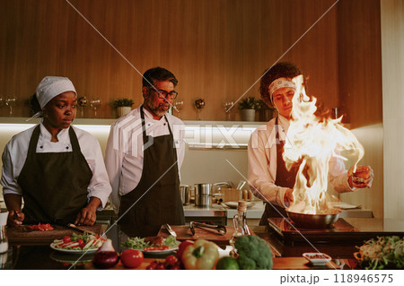 Young sous chef demonstrating frying skills while his coworkers watching process 118946575