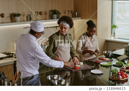 High angle shot of unrecognizable chef telling about cutting details while curly haired cook listening to him and cutting cheese, African American colleague preparing vegetables High angle shot of unrecognizable chef telling about cutting details while curly haired cook listening to him and cutting cheese, African American colleague preparing vegetables 118946615