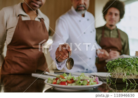 Medium close up of chefs hand holding spoon and adding olive oil into salad while his assistants standing next to him 118946630