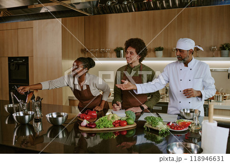 African American female intern helping her Indian master with kitchenware while her mate writing down recipe 118946631