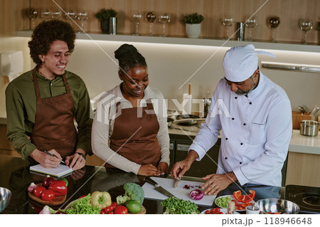 High angle shot of chef in white uniform cutting red onion while his biracial interns writing down his actions High angle shot of chef in white uniform cutting red onion while his biracial interns writing down his actions 118946648