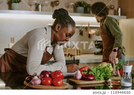 African American female cook surrounded with fresh vegetables writing down new recipe while her colleague choosing knife 118946680