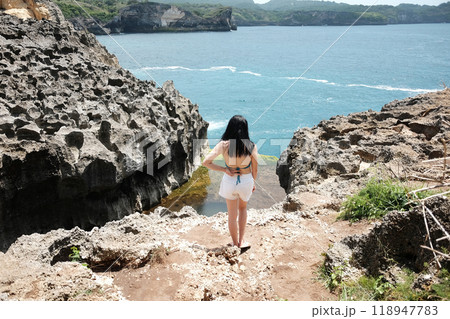 Asian woman wearing bikini with white trouser shorts is standing with her back on the rocky cliff at the sea for happy and destination travel in Bali island at Indonesia 118947783