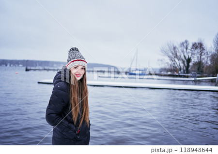 beautiful young girl with long blond hair in a hat and a long down jacket on the pier of a winter lake beautiful young girl with long blond hair in a hat and a long down jacket on the pier of a winter lake 118948644