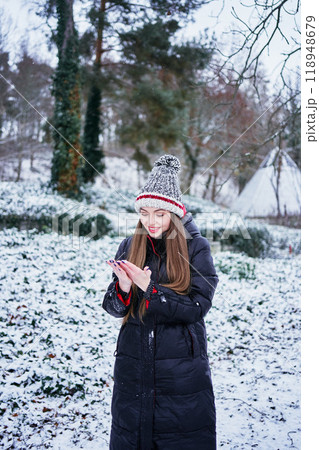a beautiful young girl with long blond hair in a hat and a long down jacket plays with snow and enjoys winter nature a beautiful young girl with long blond hair in a hat and a long down jacket plays with snow and enjoys winter nature 118948679