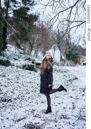 a beautiful young girl with long blond hair in a hat and a long down jacket plays with snow and enjoys winter nature 118948681