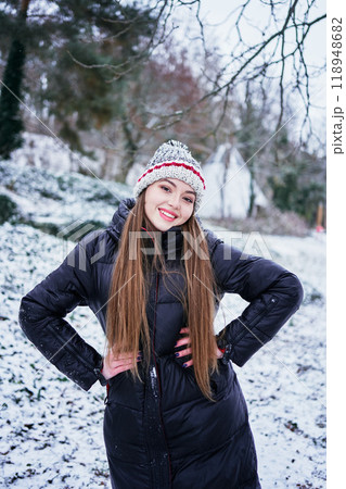 a beautiful young girl with long blond hair in a hat and a long down jacket plays with snow and enjoys winter nature 118948682