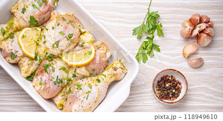 Raw uncooked chicken legs marinated with seasonings, herbs, lemon in white ceramic casserole top view on light wooden rustic background. Preparing healthy meal with marinated chicken drumsticks 118949610