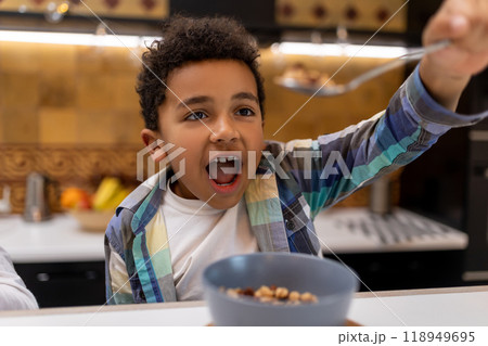 Dark-skinned boy sitting at the table and eating cereals Dark-skinned boy sitting at the table and eating cereals 118949695
