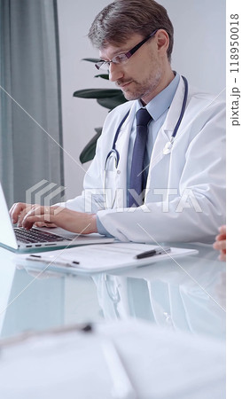 Male physician in white coat focused on laptop data in a modern clinic office while analyzing patient information on laptop. Medicine concept 118950018