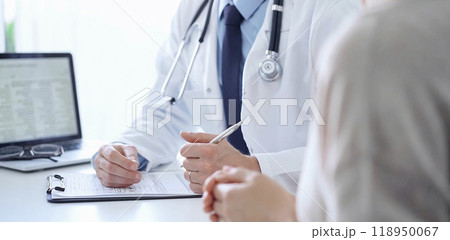 A doctor and a patient. The physician, wearing a white medical coat over a blue shirt and tie, is filling out a medical record form during a consultation in the clinic. Medical service A doctor and a patient. The physician, wearing a white medical coat over a blue shirt and tie, is filling out a medical record form during a consultation in the clinic. Medical service 118950067