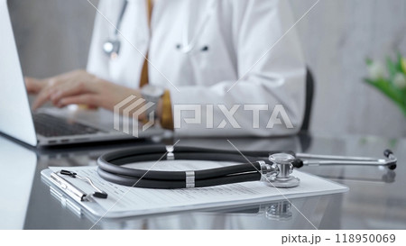 Stethoscope and clipboard with medication history records are on a glass desk behind a physician using a laptop computer. Medicine concept Stethoscope and clipboard with medication history records are on a glass desk behind a physician using a laptop computer. Medicine concept 118950069