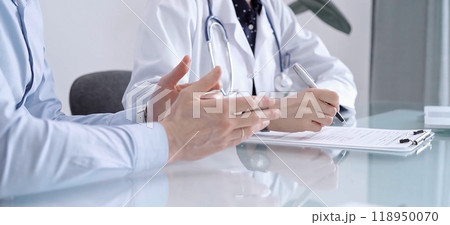 Doctor consulting patient in clinic. Female physician is writing notes while talking with a man wearing blue shirt, close up. Medicine concept Doctor consulting patient in clinic. Female physician is writing notes while talking with a man wearing blue shirt, close up. Medicine concept 118950070