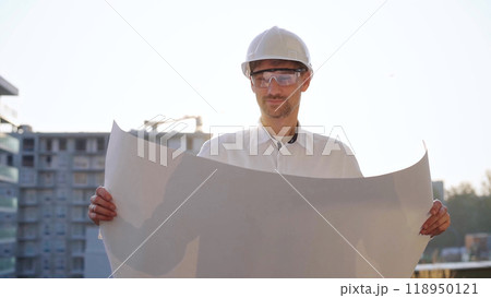 Male construction site manager, wearing safety helmets and glasses, is holding blueprints and inspecting a building site during sunrise or sunset, front side view. Architecture and engineering Male construction site manager, wearing safety helmets and glasses, is holding blueprints and inspecting a building site during sunrise or sunset, front side view. Architecture and engineering 118950121