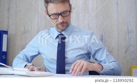 Male accountant with blue t-shirt concentrating while calculating costs and taking notes at his desk. Taxes, audit in business Male accountant with blue t-shirt concentrating while calculating costs and taking notes at his desk. Taxes, audit in business 118950292