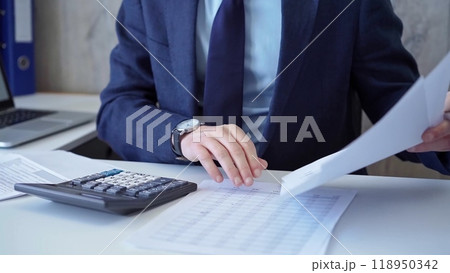 Businessman accountant with blue suit is calculating numbers for a financial audit and taking notes with a silver pen. Taxes and audit concept 118950342