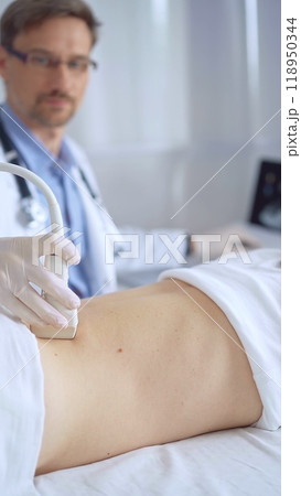 Doctor man wearing white protective gloves is using ultrasound equipment on the back of a patient, woman. The results are on a monitor in the background. Medicine service Doctor man wearing white protective gloves is using ultrasound equipment on the back of a patient, woman. The results are on a monitor in the background. Medicine service 118950344