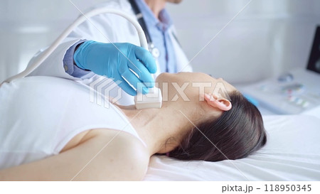 Doctor man wearing blue medical gloves is using ultrasound equipment on neck of female patient lying down for a medical examination 118950345