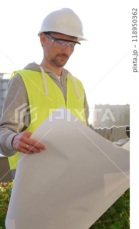 Man an architect or engineer wearing safety hard hat and vest holding blueprint and looking to construction site at sunrise, front vertical view. Architecture concept Man an architect or engineer wearing safety hard hat and vest holding blueprint and looking to construction site at sunrise, front vertical view. Architecture concept 118950362