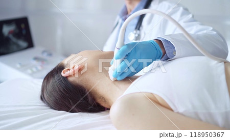 Doctor man wearing blue medical gloves is using ultrasound equipment on neck of female patient lying down for a medical examination 118950367