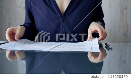 Businessperson woman in dark blue blouse analyzing document at glass desk. Close-up of a professional auditor or lawyer reviewing a lengthy paper report in office setting. Business people concept 118950379