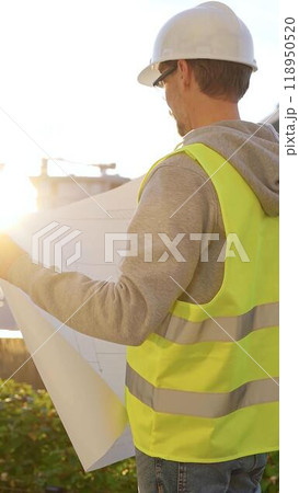 Man an architect or engineer wearing safety hard hat and vest holding blueprint and looking to construction site at sunrise, back vertical view. Architecture and engineering Man an architect or engineer wearing safety hard hat and vest holding blueprint and looking to construction site at sunrise, back vertical view. Architecture and engineering 118950520